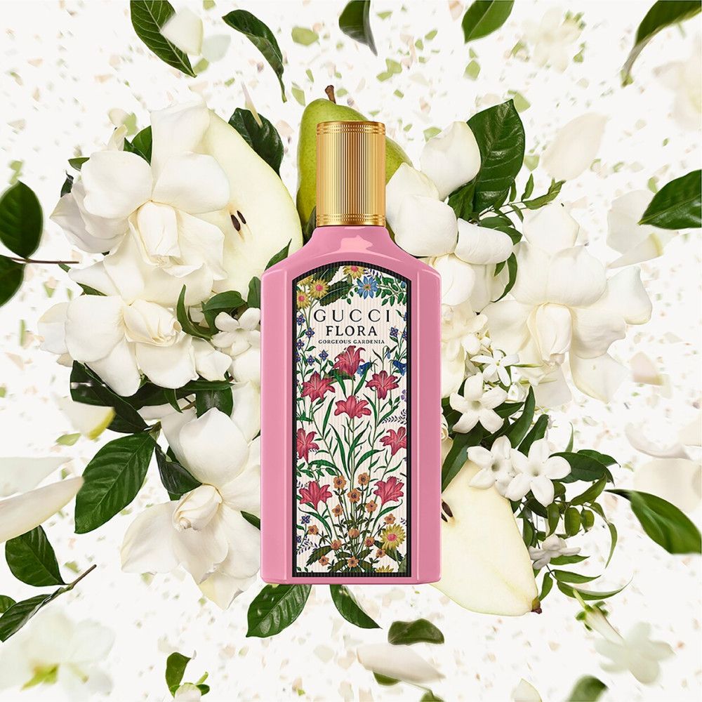 Flacon rose avec bouchon doré, entouré de fleurs blanches et feuilles vertes. Texte Gucci Flora.