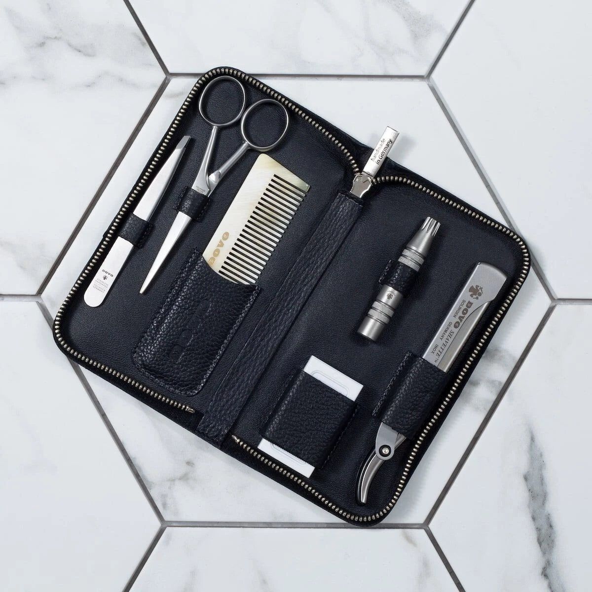 Zwarte etui met rits met baardverzorgingsgereedschap: schaar, kam, pincet, neushaartrimmer, nagelknipper en etui. Op tegels.