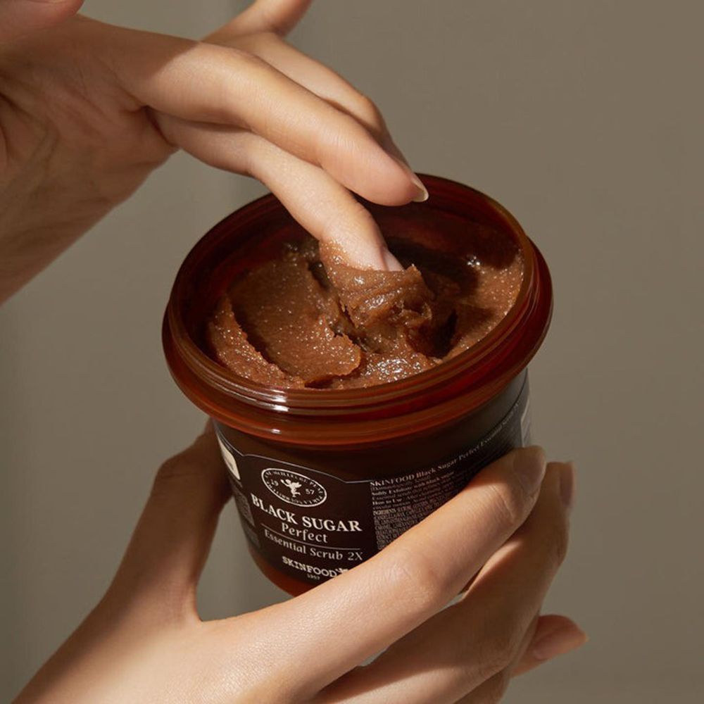 Handen houden een open bruine pot vast. Vingers raken de scrub aan. Opschrift: Black Sugar Perfect Essential Scrub 2X.