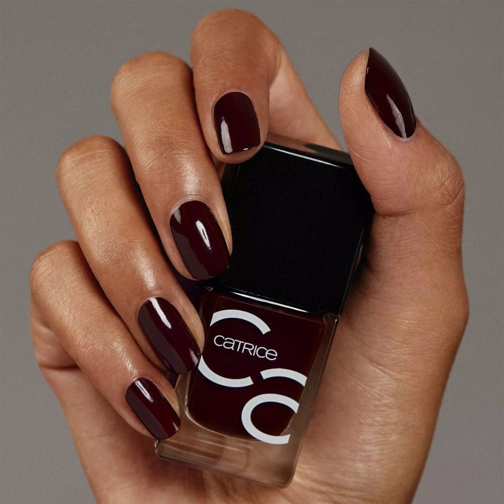 Main avec ongles vernis et flacon de vernis à ongles. Ongles bordeaux, flacon avec logo Catrice.