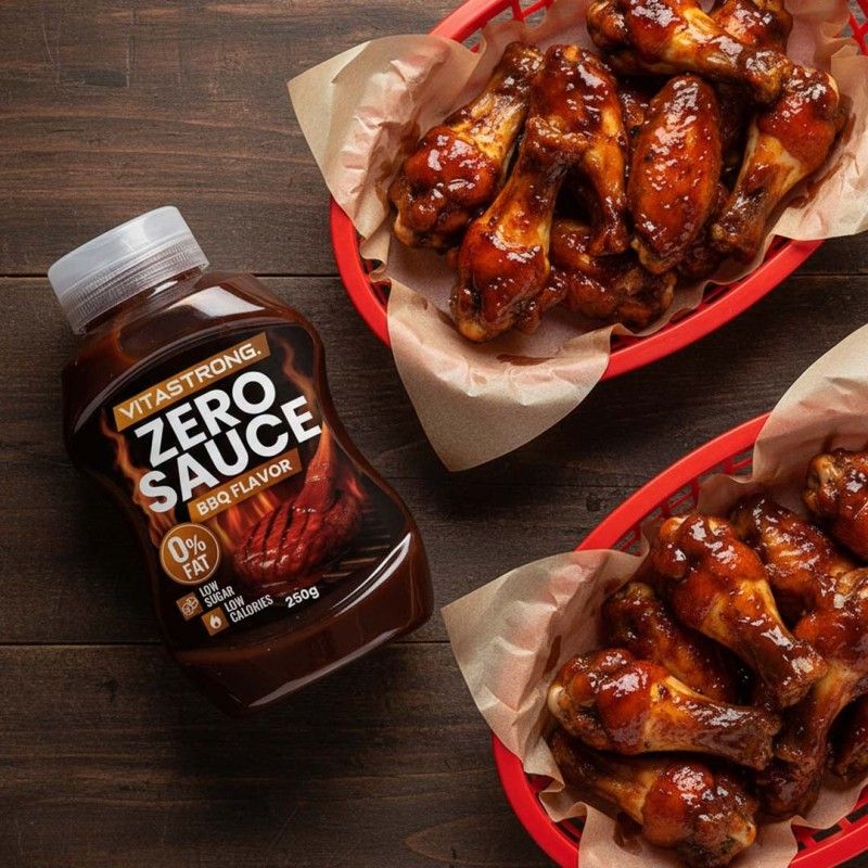 Bruine fles met witte dop. Opschrift: Vitastrong Zero Sauce BBQ Flavor. 0% vet, weinig suiker, weinig calorieën. 250g. Naast twee manden met kippenvleugels.