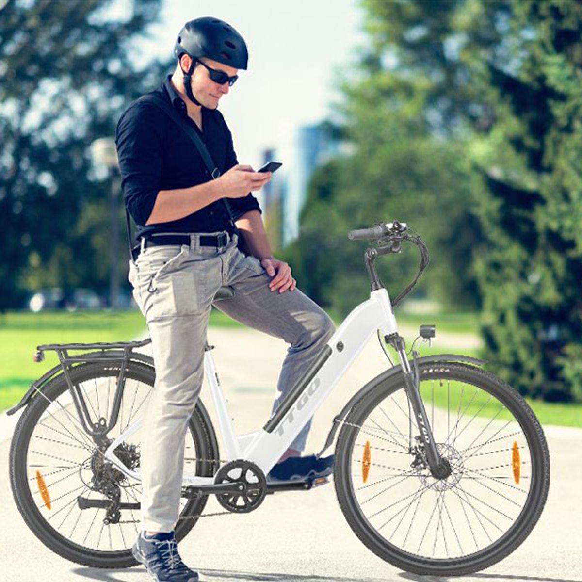 Homme debout à côté d'un vélo électrique blanc. Casque, lunettes de soleil et smartphone. Arrière-plan : arbres et bâtiments.