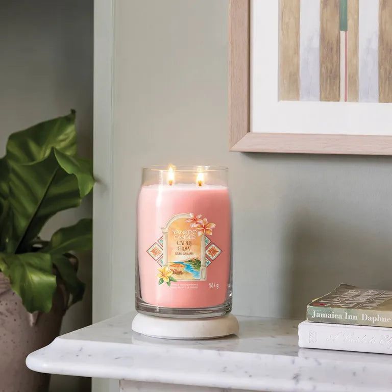 Bougie rose dans un verre, deux mèches, sur socle blanc. Inscription : Capri Glow. Éléments décoratifs.