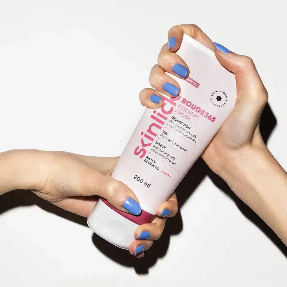 Handen houden crèmetube SKINLICK Rouge365 Essential Cream. Roze-wit ontwerp, rode streep. Blauwe nagels. Tekst zichtbaar.