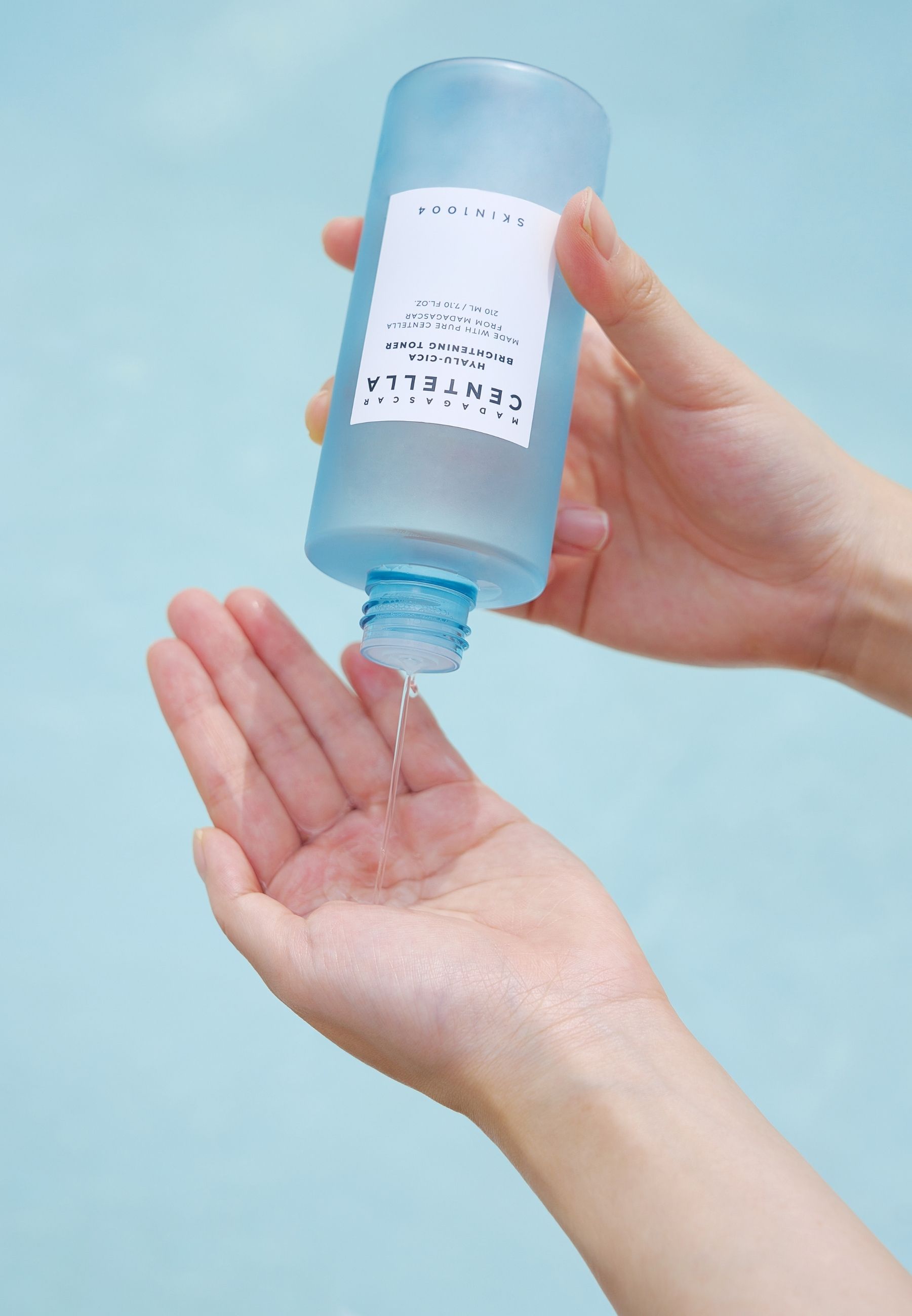 Handen houden een lichtblauwe fles vast. Vloeistof stroomt eruit. Opschrift: MADAGASCAR CENTELLA. SKIN1004.