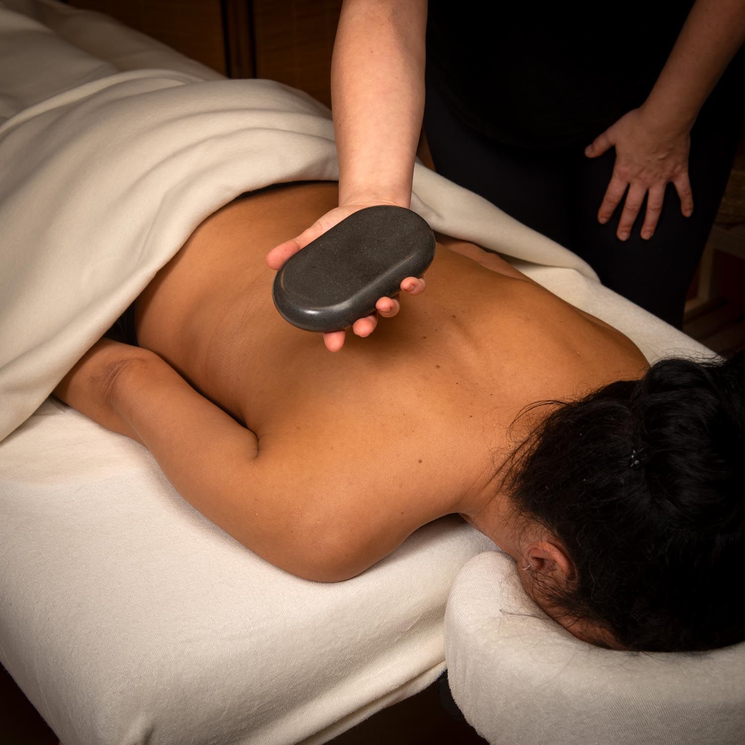 Grosse pierre ovale sur le dos d'une personne allongée. La personne est sur une table de massage, recouverte d'un drap blanc.