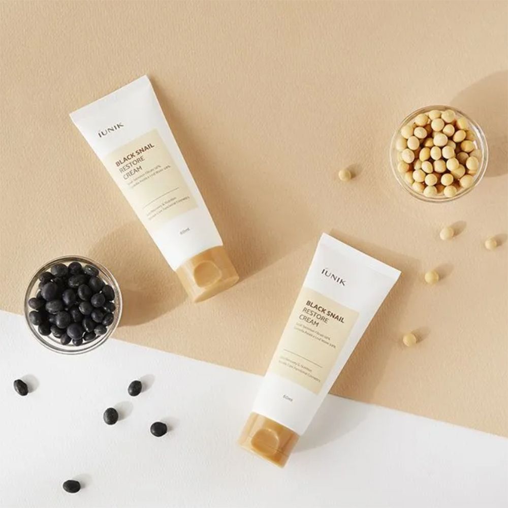 Deux tubes de crème avec informations produit, à côté de bols de haricots. Inscription: Black Snail Restore Cream. Fond beige.