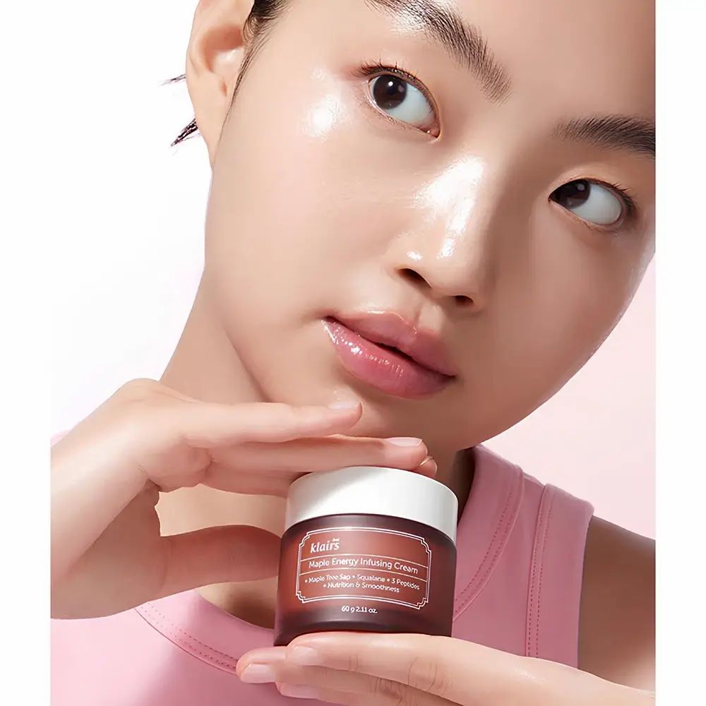 Vrouw houdt bruine crèmepot vast. Opschrift: Dear Klairs Maple Energy Infusing Cream. Gezicht glanst. Roze top.