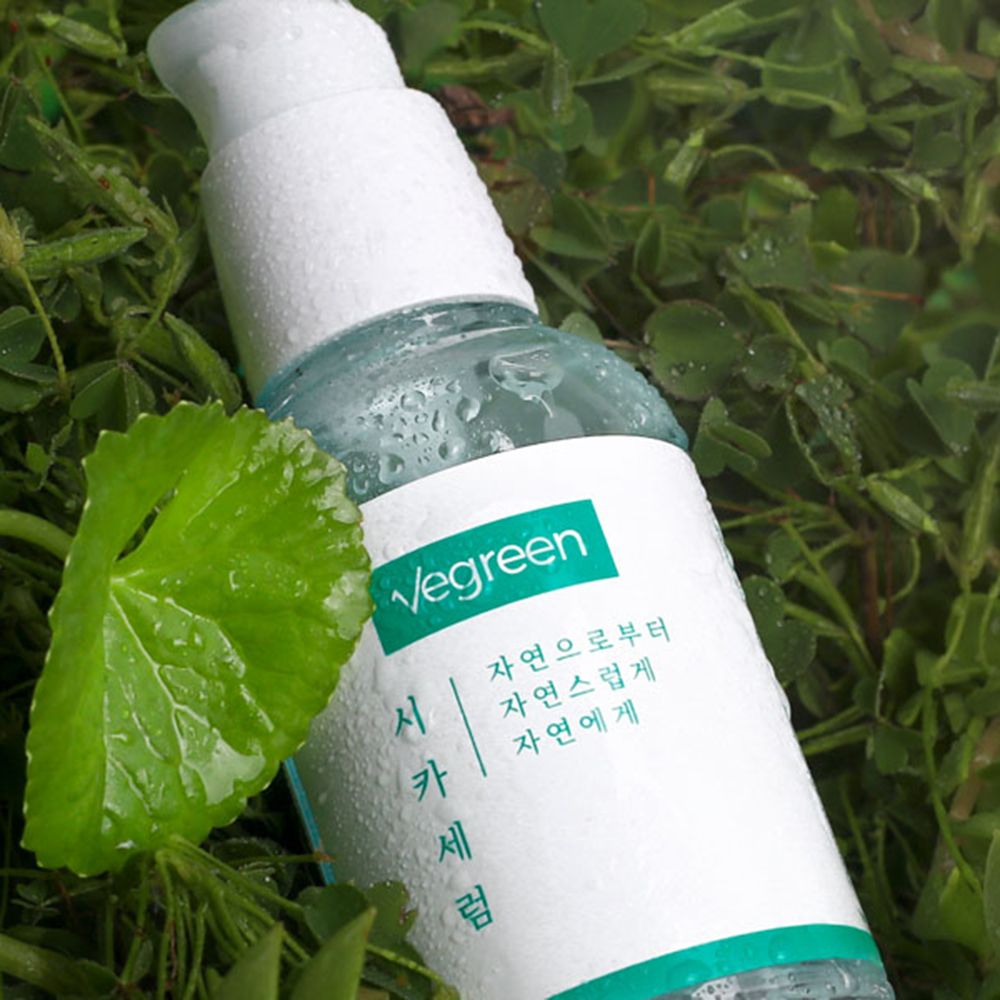 Gezichtsserumfles in het gras. Witte pompdop, groen etiket met tekst. Druppels op de fles en een blad.