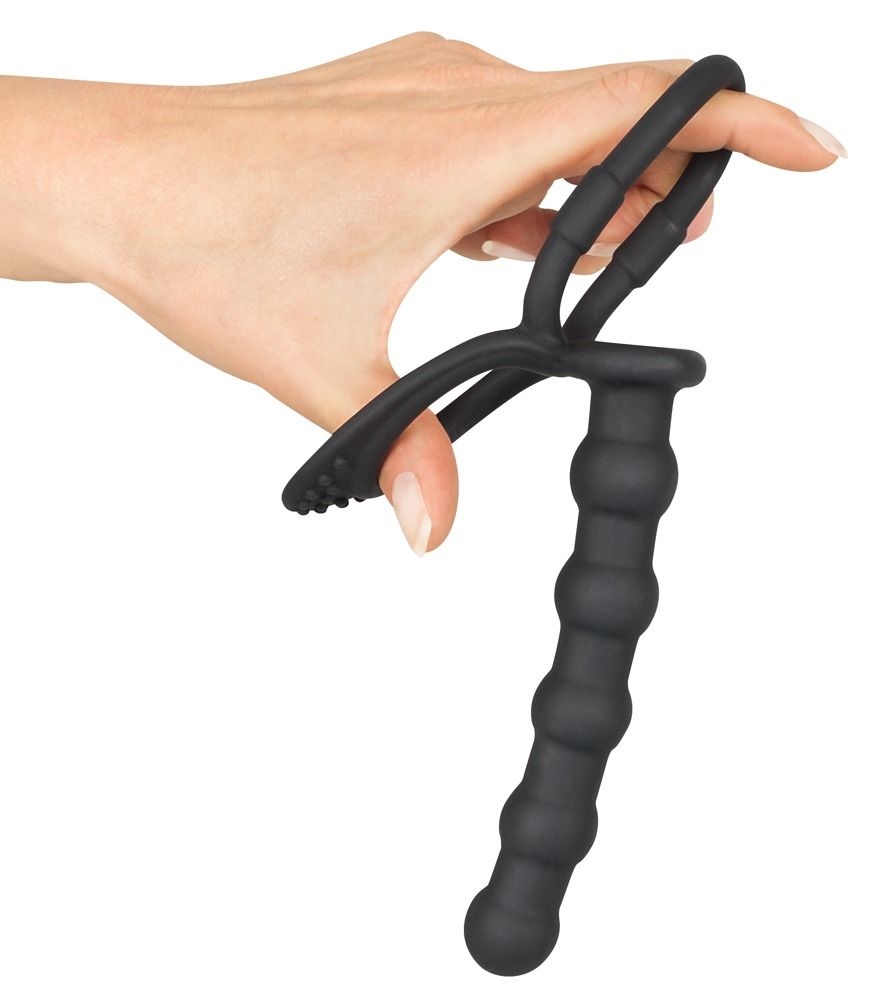 Hand houdt zwarte penis- en ballenring met anale dildo vast. Ring met nopjes, dildo met ribbels.