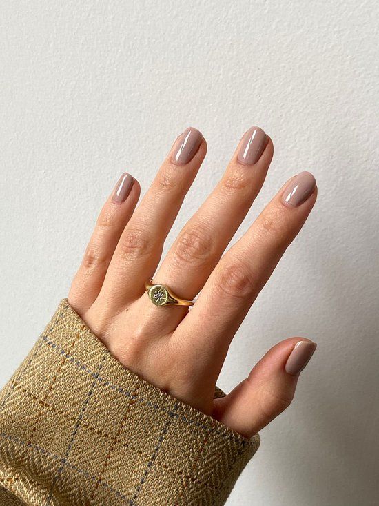 Main avec ongles vernis dans une teinte beige clair. Bague dorée avec pierre. Fond de couleur peau.