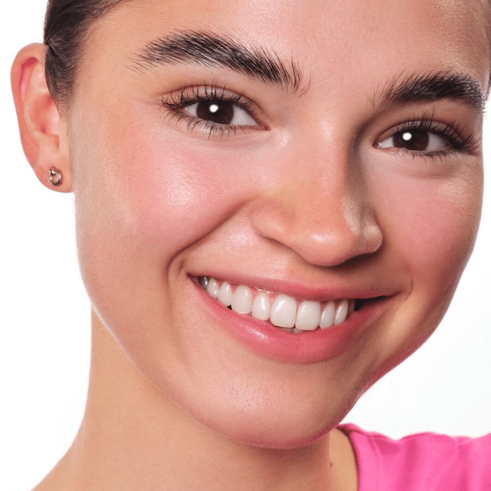 Gros plan d'un visage souriant. Teint, sourcils, yeux, dents. Porte des boucles d'oreilles.
