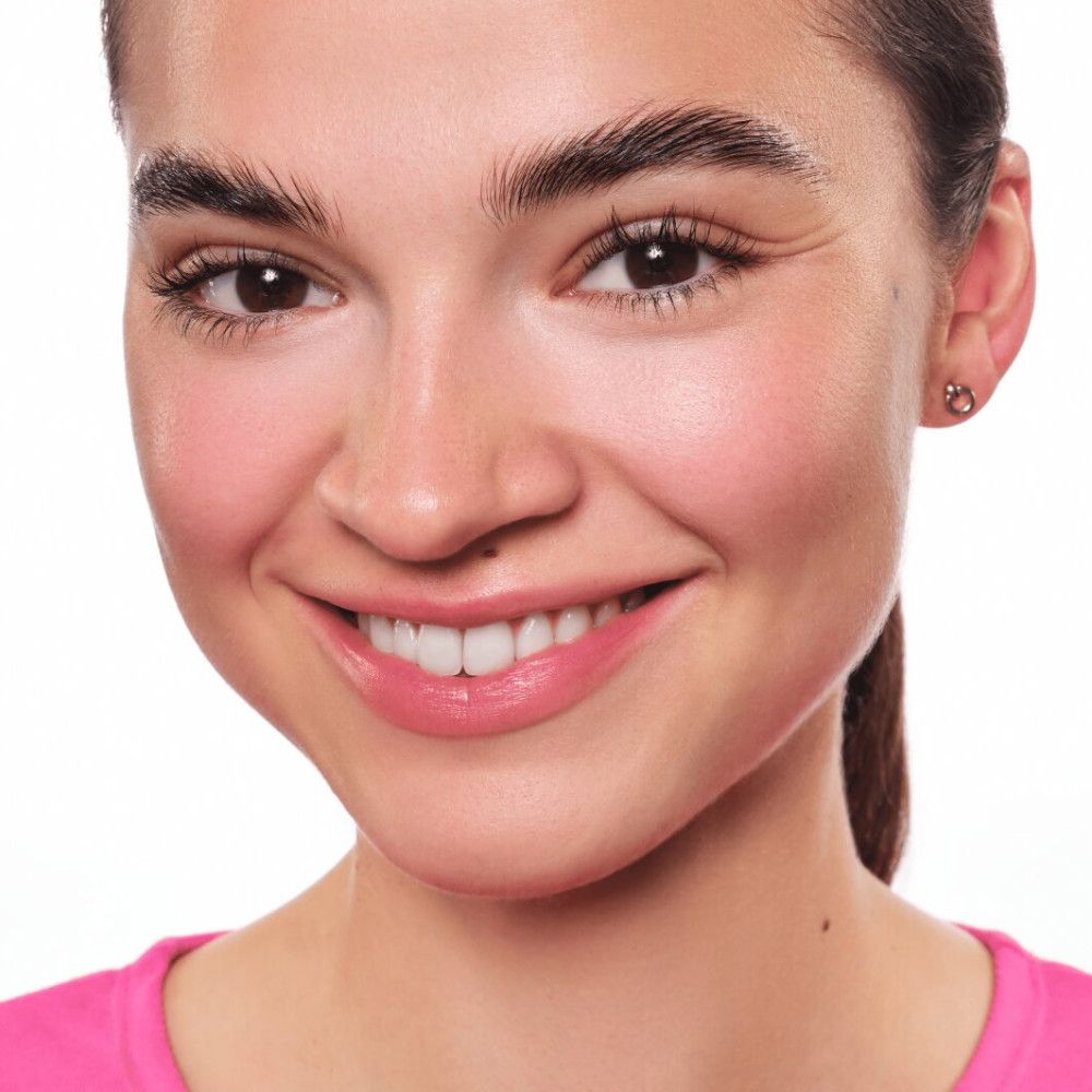 Portret van een lachende vrouw met gebruinde huid. Ze draagt een roze top. Duidelijke wenkbrauwen en lippenstift.