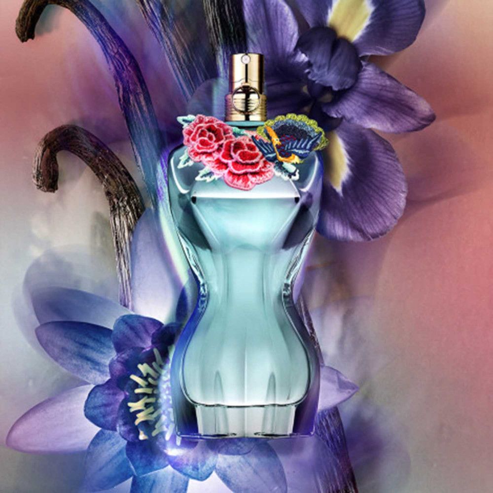 Flacon de parfum bleu en forme de corps féminin, décoré de fleurs. Fermeture dorée. Entouré de fleurs violettes et roses.