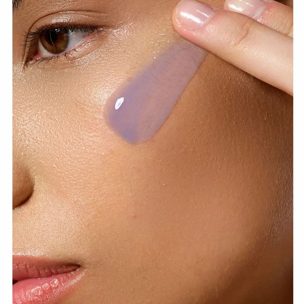 Close-up van een gezicht. Een hand brengt serum aan op de huid. Serum is paars.