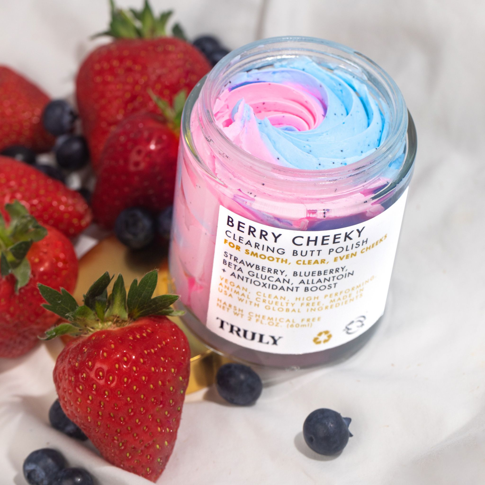 Glazen pot met roze en blauw product, omringd door aardbeien en bosbessen. Productlabel zichtbaar.