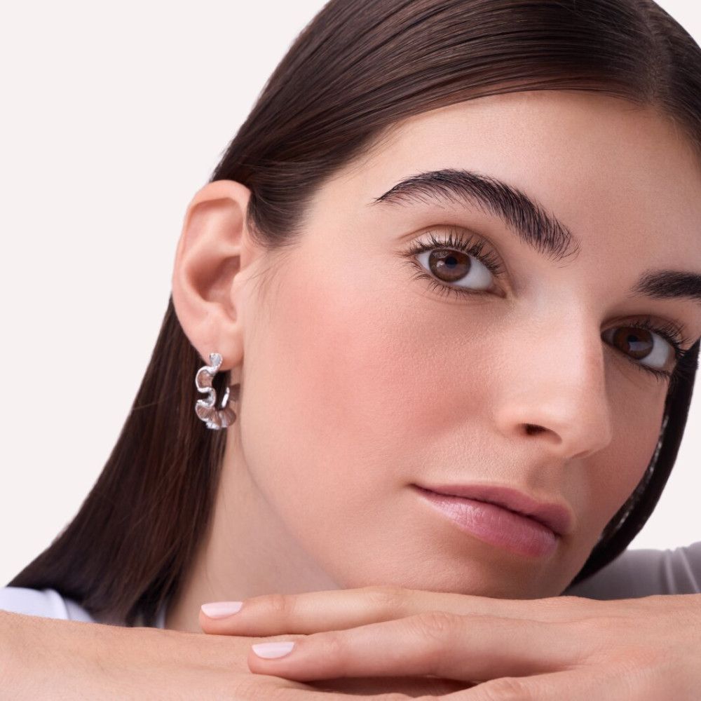 Portret van een vrouw met roze blush op de wangen. Ze steunt haar kin op haar handen. Lichte huid, bruine ogen, donker haar.