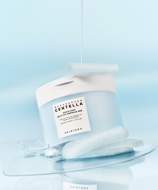 Contenant bleu et blanc, liquide qui coule. Pads et liquide sur une surface. Texte: CENTELLA, HYALU-CICA JELLY-FIT AMPULE PAD, SKIN1004.