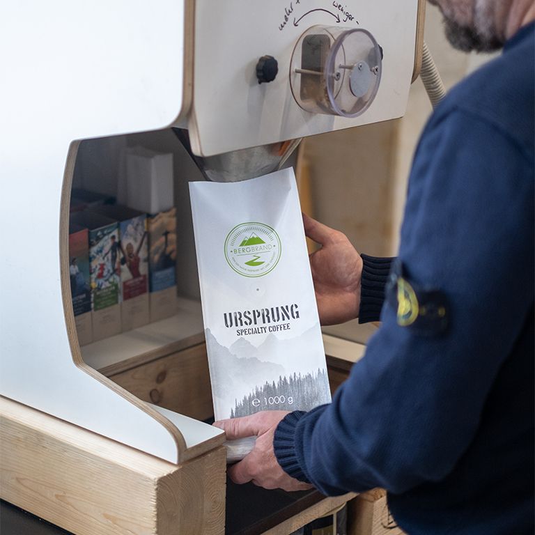 Koffiebonenverpakking. Wit, met logo en tekst. Persoon houdt de verpakking vast. 1000g.
