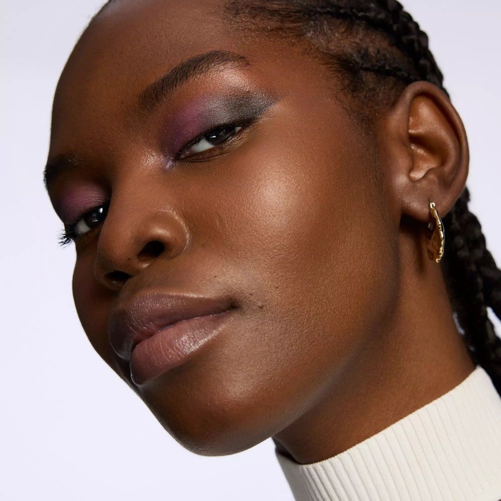 Gros plan d'une femme à la peau foncée portant du fard à paupières violet. Elle a des cheveux tressés et porte une boucle d'oreille dorée.