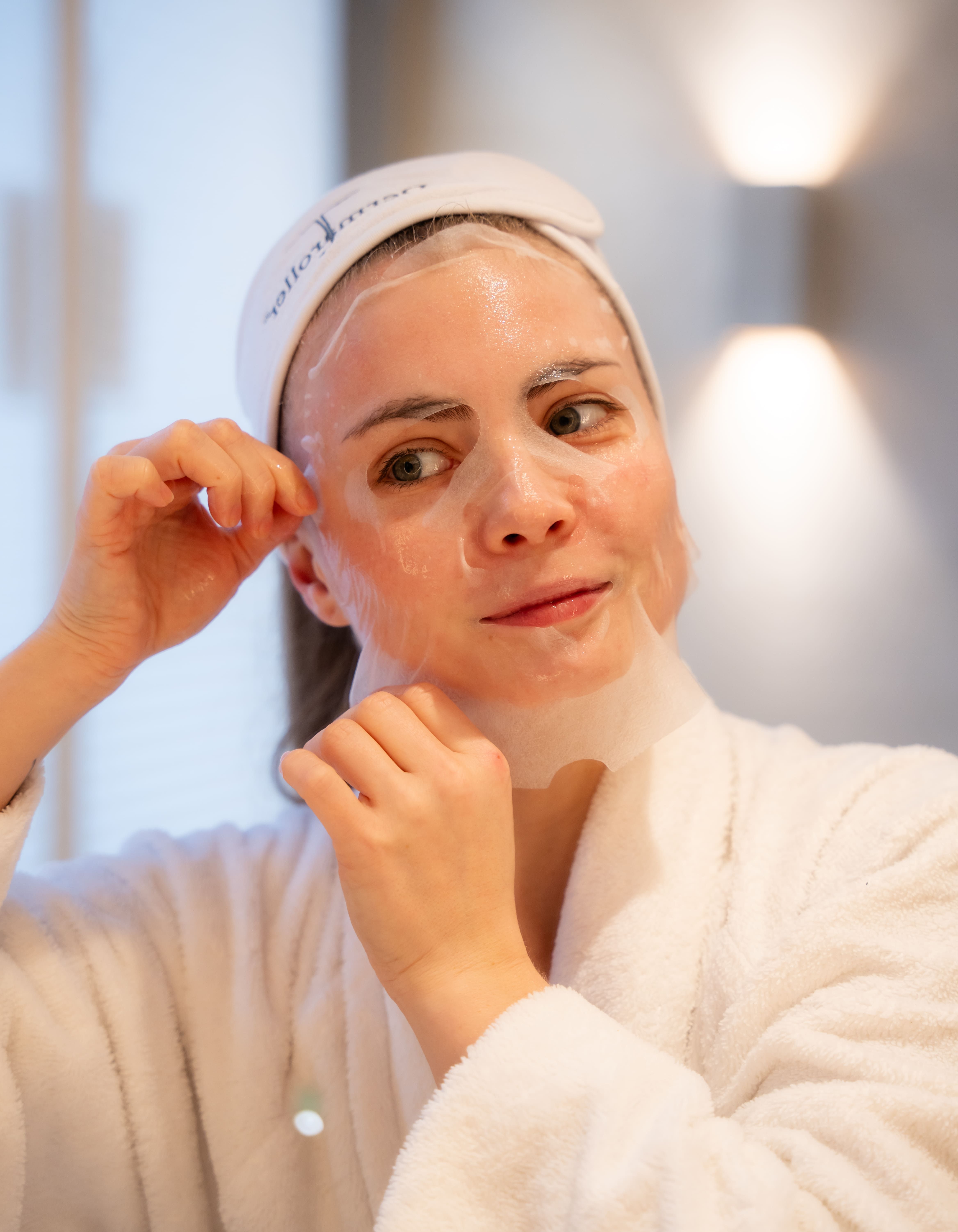 Vrouw met witte hoofdband en badjas brengt een sheetmasker aan.