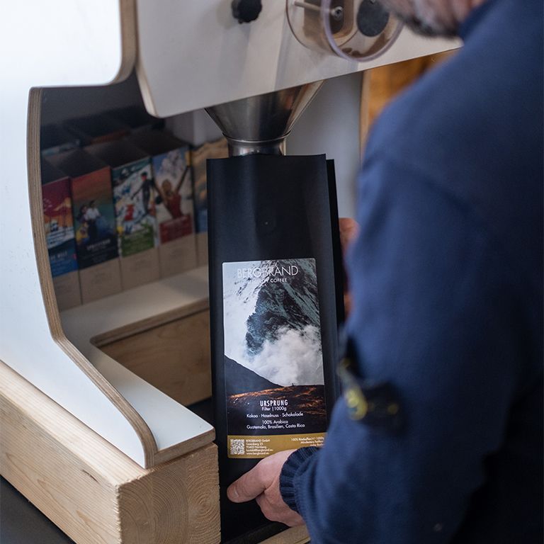 Personne tenant un paquet de café noir. Texte : BERGBRAND, Ursprung, Filter, 250g. Ingrédients : cacao, noisette, chocolat, 100% Arabica.