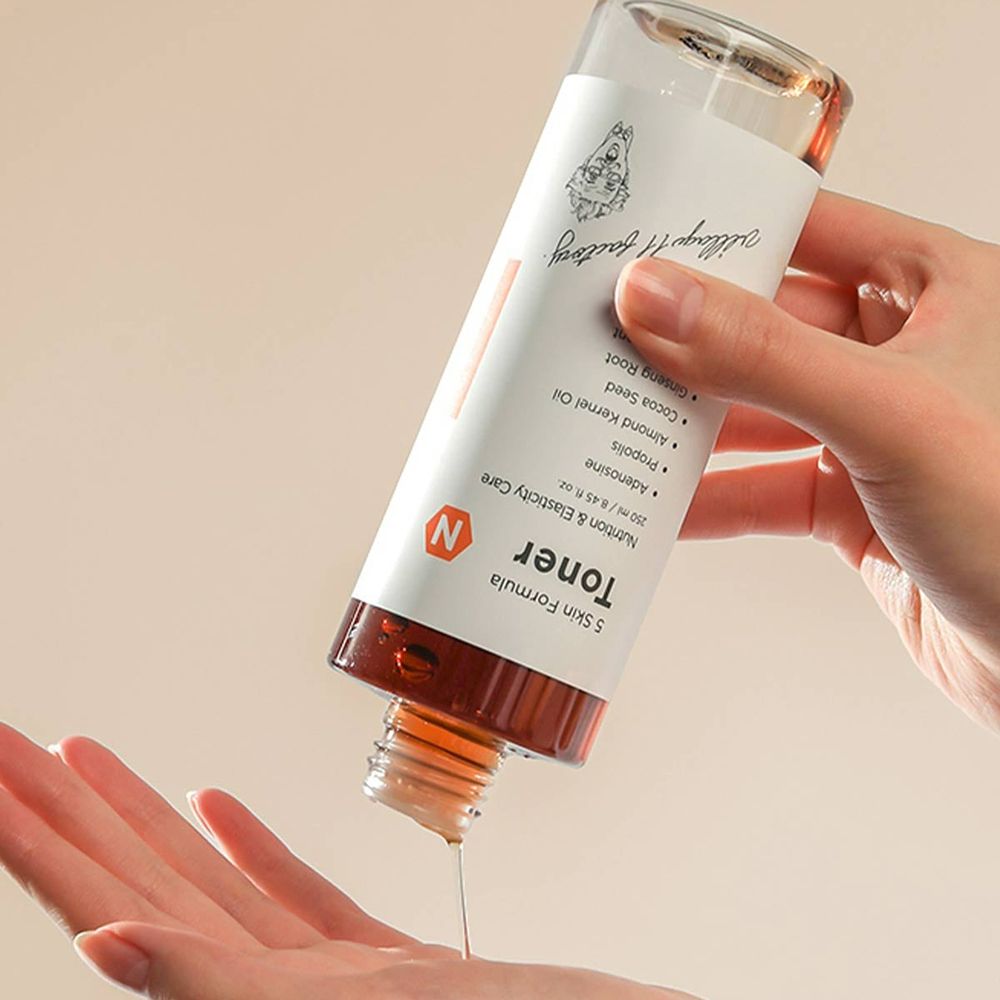 Fles wordt boven een hand gehouden. Vloeistof druppelt eruit. Label met tekst: Toner, 5 Skin Formula, N. Oranje streep.
