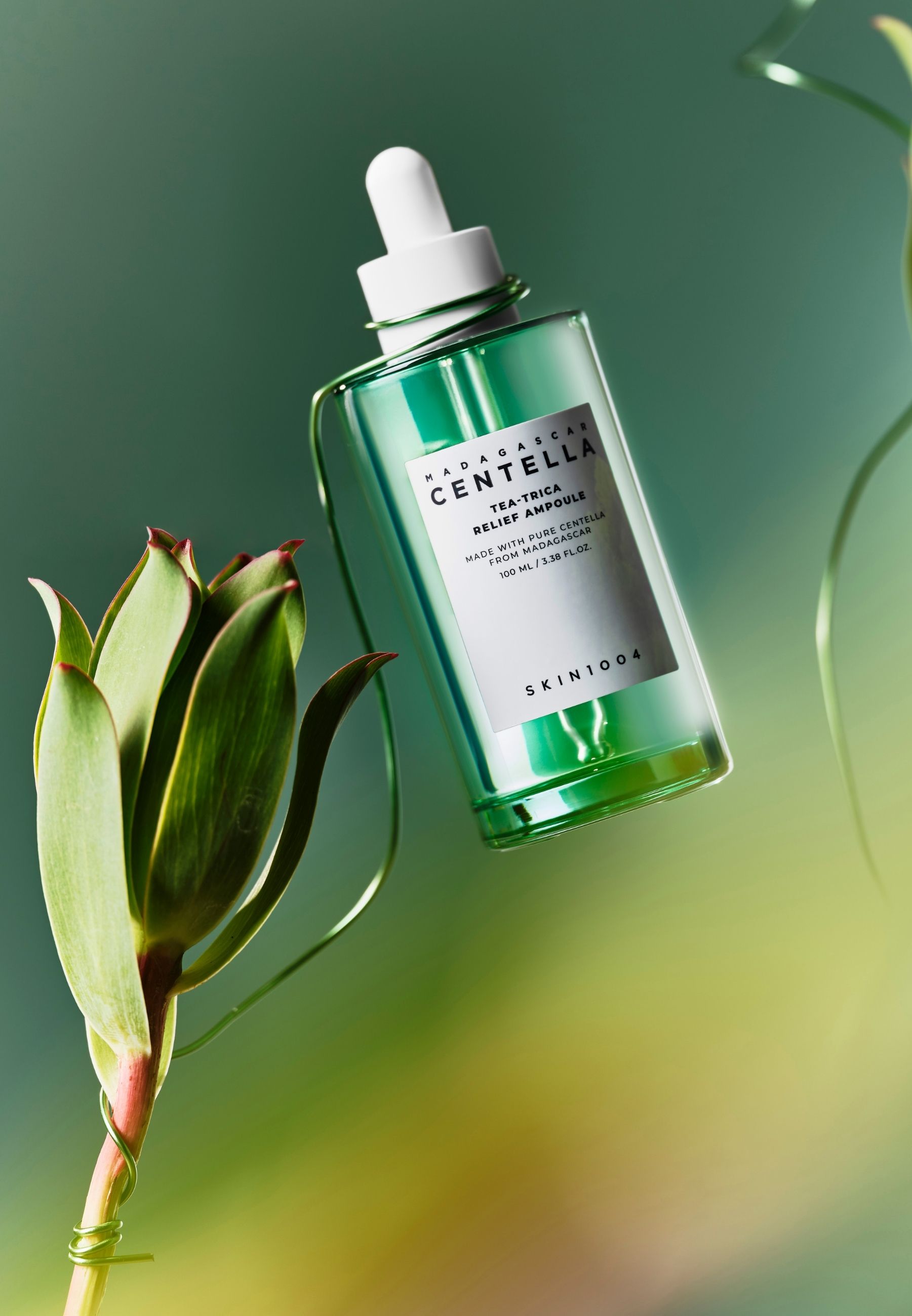 Groene glazen fles met pipet, omringd door groene planten. SKIN1004 logo zichtbaar.