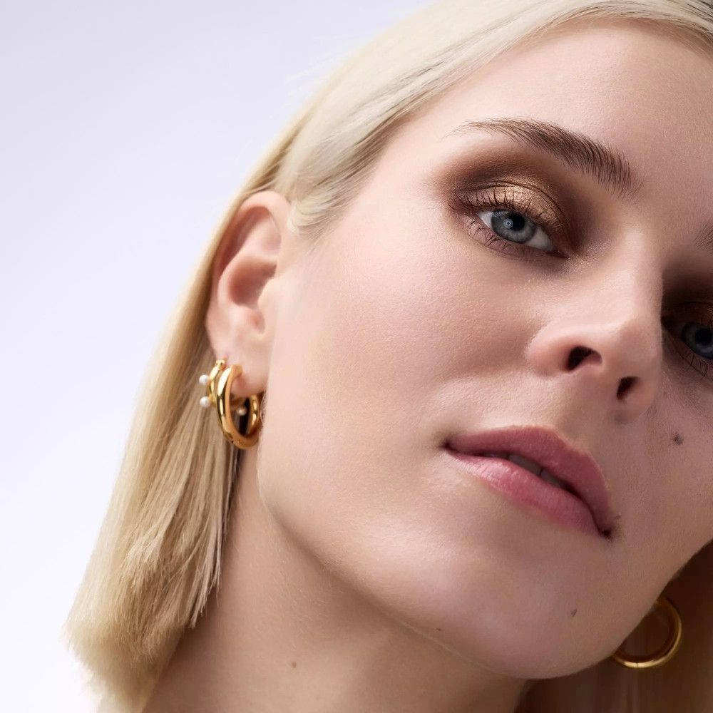 Gros plan d'un visage avec des cheveux blonds et des créoles dorées. De l'ombre à paupières est appliquée sur les paupières.