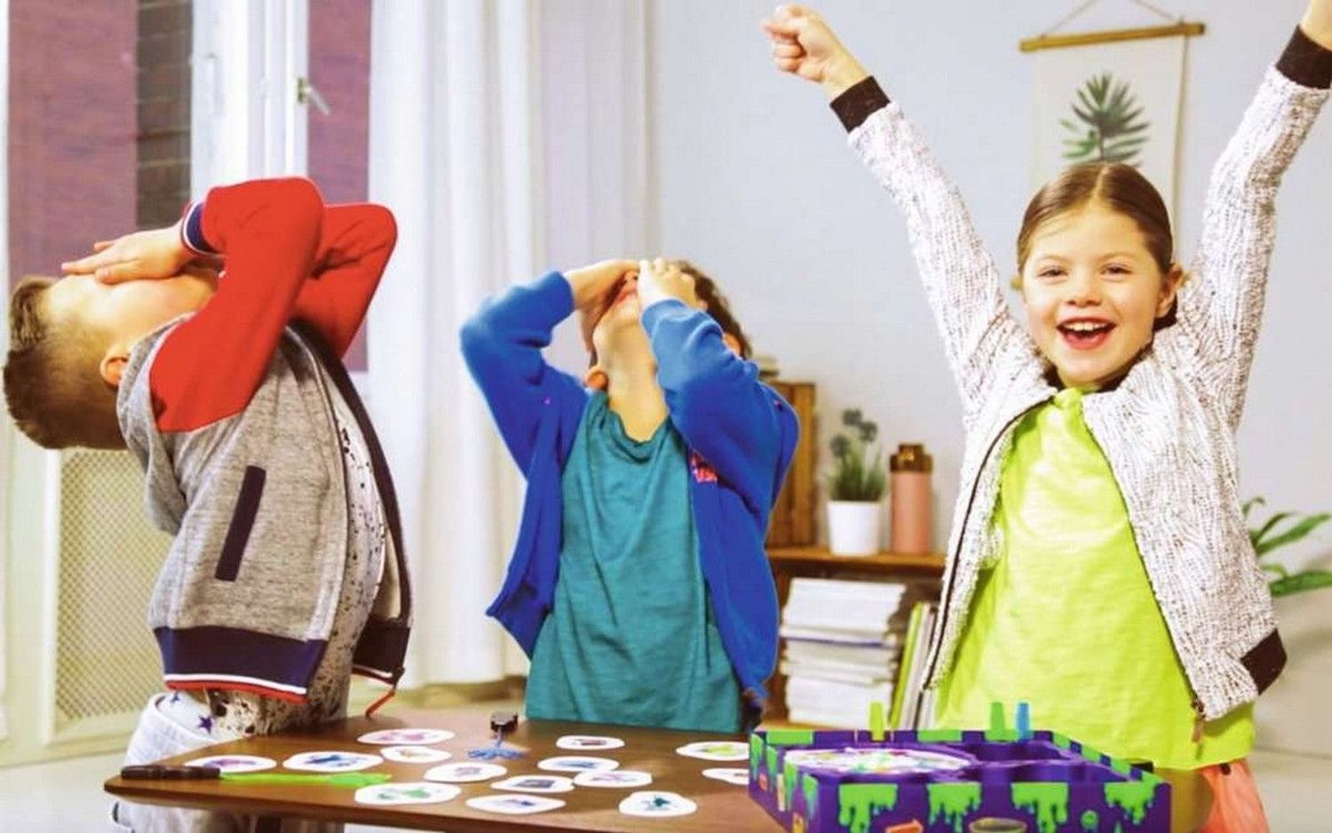 Drie kinderen spelen. Eén kind juicht, twee bedekken hun ogen. Bord en kaarten op tafel.