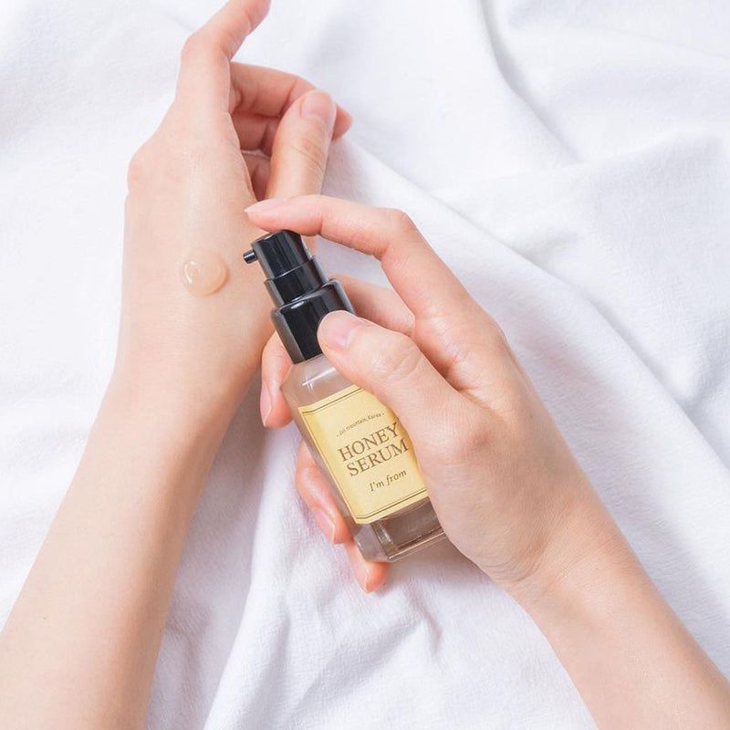 Mains tenant un flacon de sérum. Sérum sur le dos de la main. L'étiquette indique "HONEY SERUM" et "I'm from".