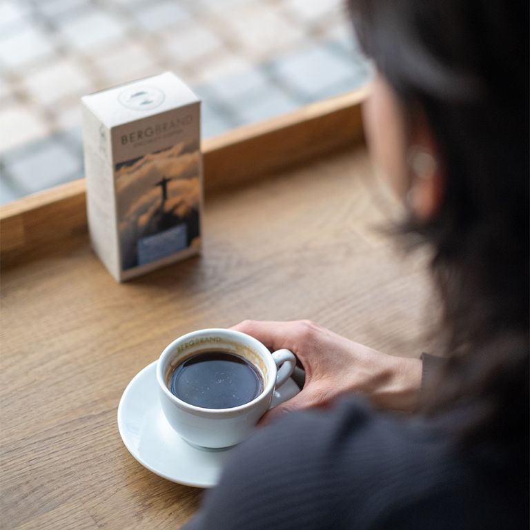 Une personne tient une tasse et une soucoupe à café. À côté, un paquet de BERGBRAND Gipfelkreuz Specialty Coffee.