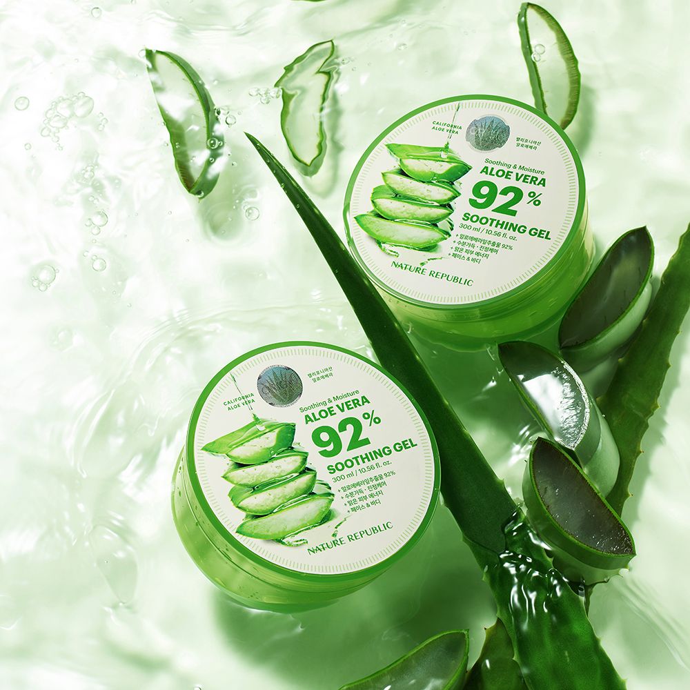 Twee groene gelpotten met deksels. Naast, aloë vera bladeren. Op de deksel staat "Aloe Vera 92% Soothing Gel".