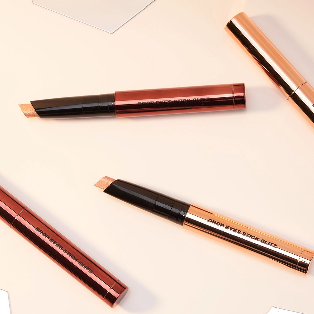 Plusieurs fards à paupières Drop Eyes Stick Glitz sont posés sur une surface claire. Les bâtons sont dorés et ont des applicateurs noirs.