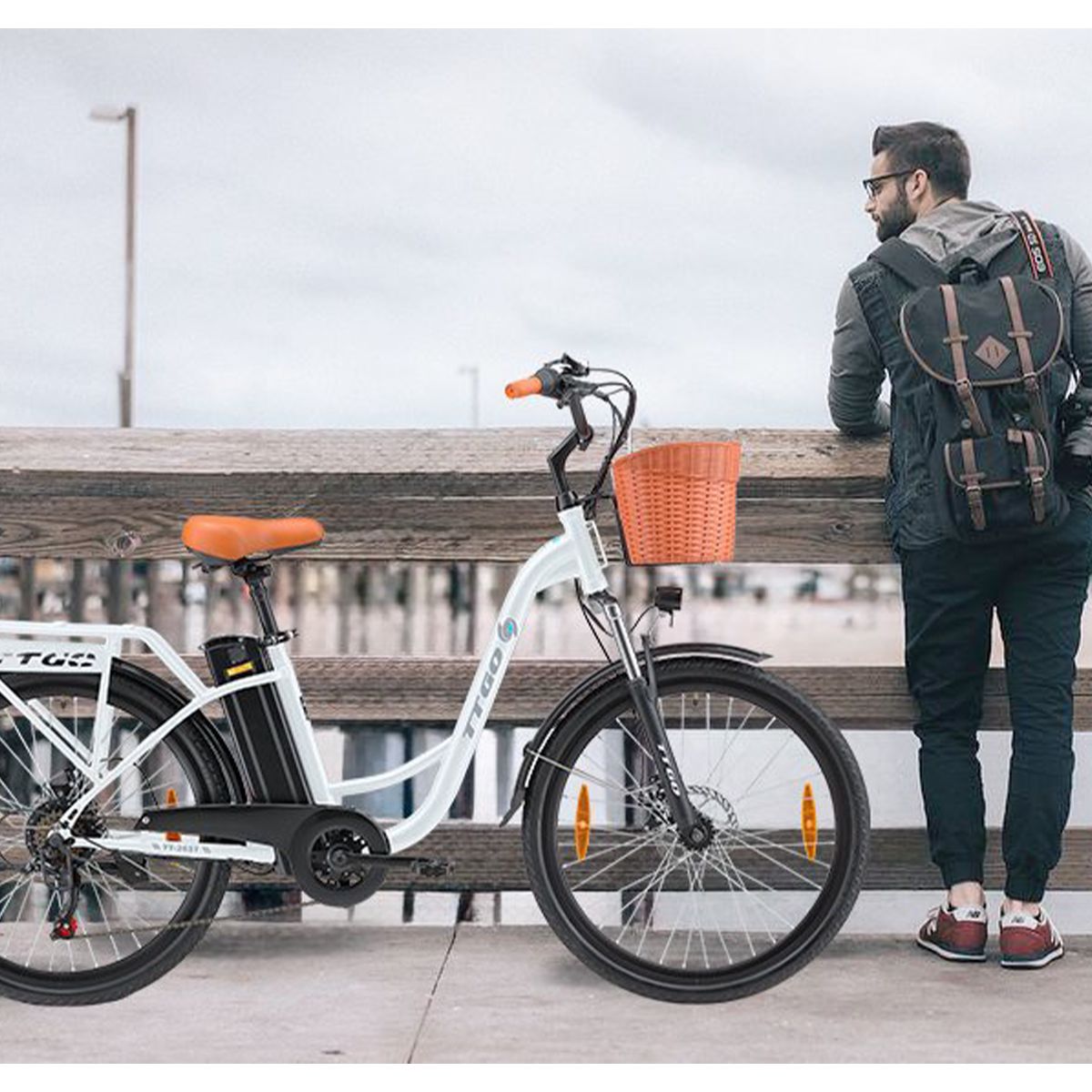 Persoon staat naast witte elektrische fiets met mand. Bruin zadel en handvatten. TTGO-logo. Achtergrond: steiger.
