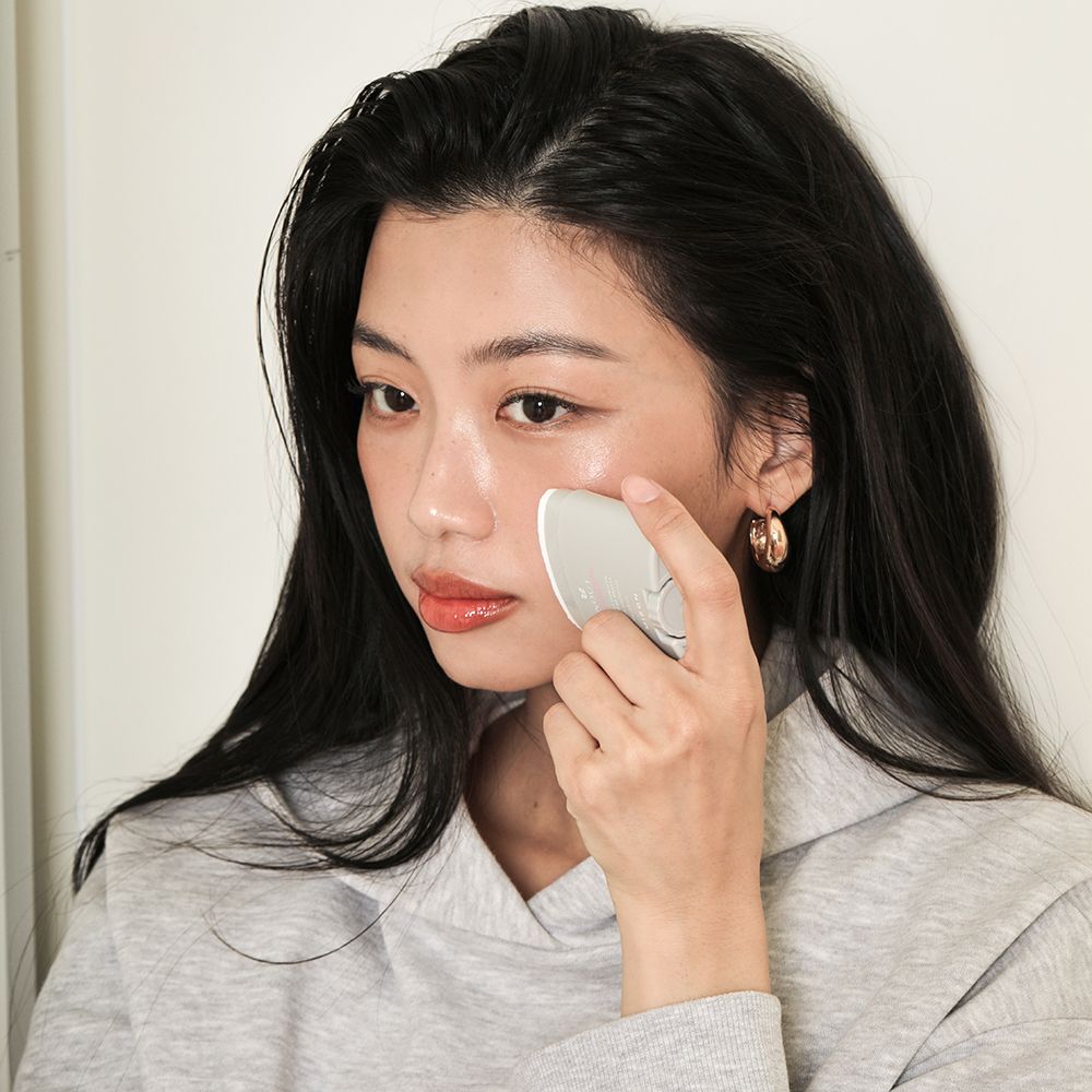 Vrouw met donker haar brengt een grijs product aan op haar gezicht. Ze draagt een grijze hoodie.