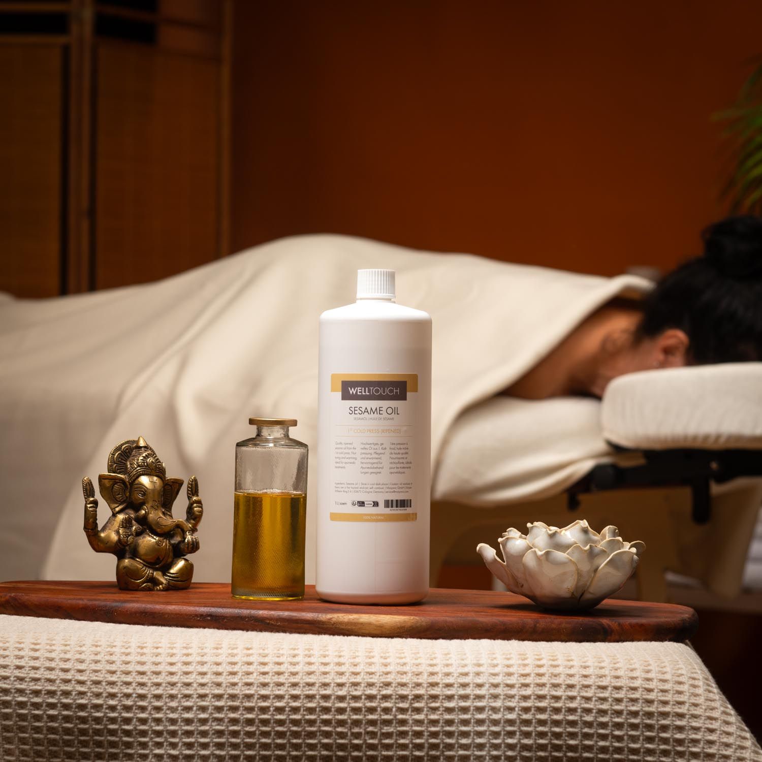 Welltouch sesamolie fles, glazen fles, goudkleurige olie, houten dienblad, Boeddha beeld, lotusbloem, massagetafel.