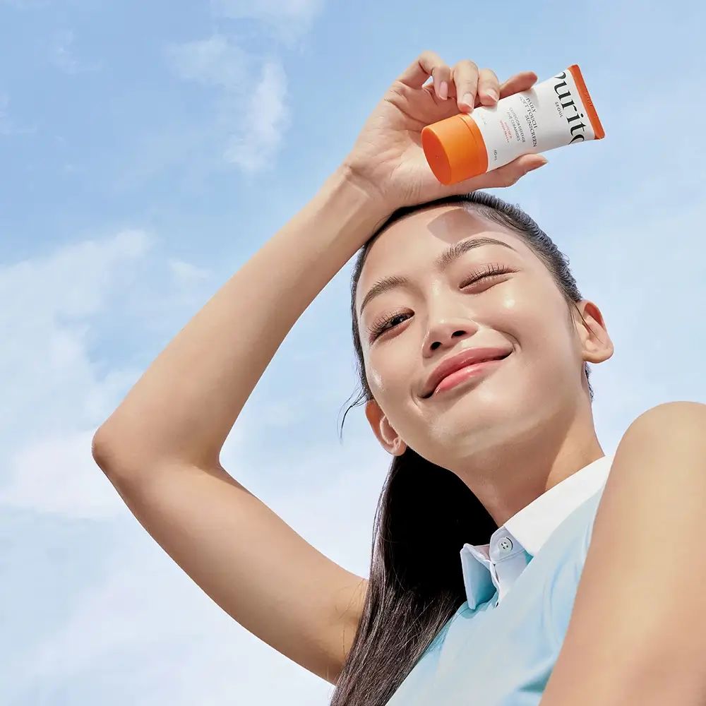 Vrouw houdt een tube zonnecrème boven haar hoofd. Blauwe lucht op de achtergrond. Product: PURITO.