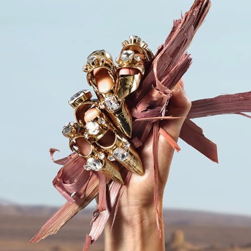 Hand met gouden ringen met edelstenen, houdt stukken hout vast. Achtergrond: landschap.