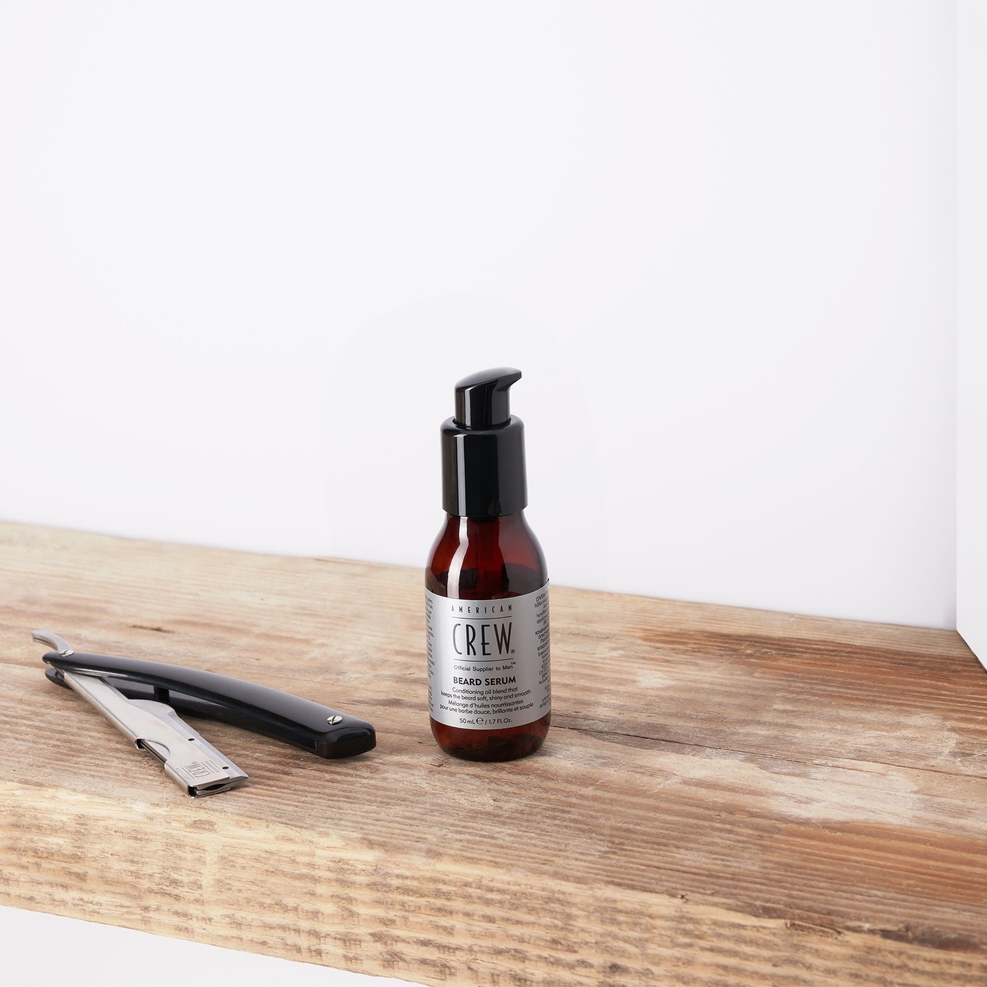 Un flacon de sérum à barbe AMERICAN CREW et un rasoir sur une planche de bois. Flacon noir avec étiquette argentée.