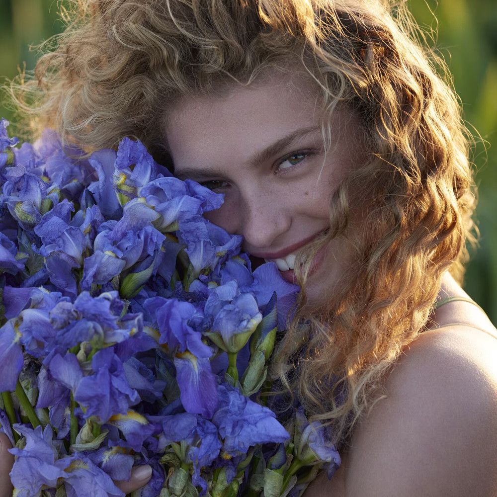 Portret van een vrouw met krullend haar die een boeket blauwe bloemen omarmt en glimlacht.