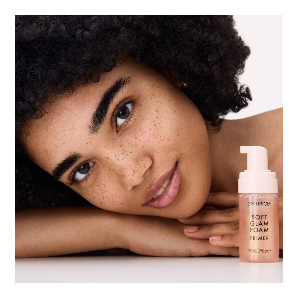 Vrouw met krullen en foundationfles. Opschrift: Catrice, Soft Glam Foam Primer, 32,5 ml/1,09 fl.oz.e.