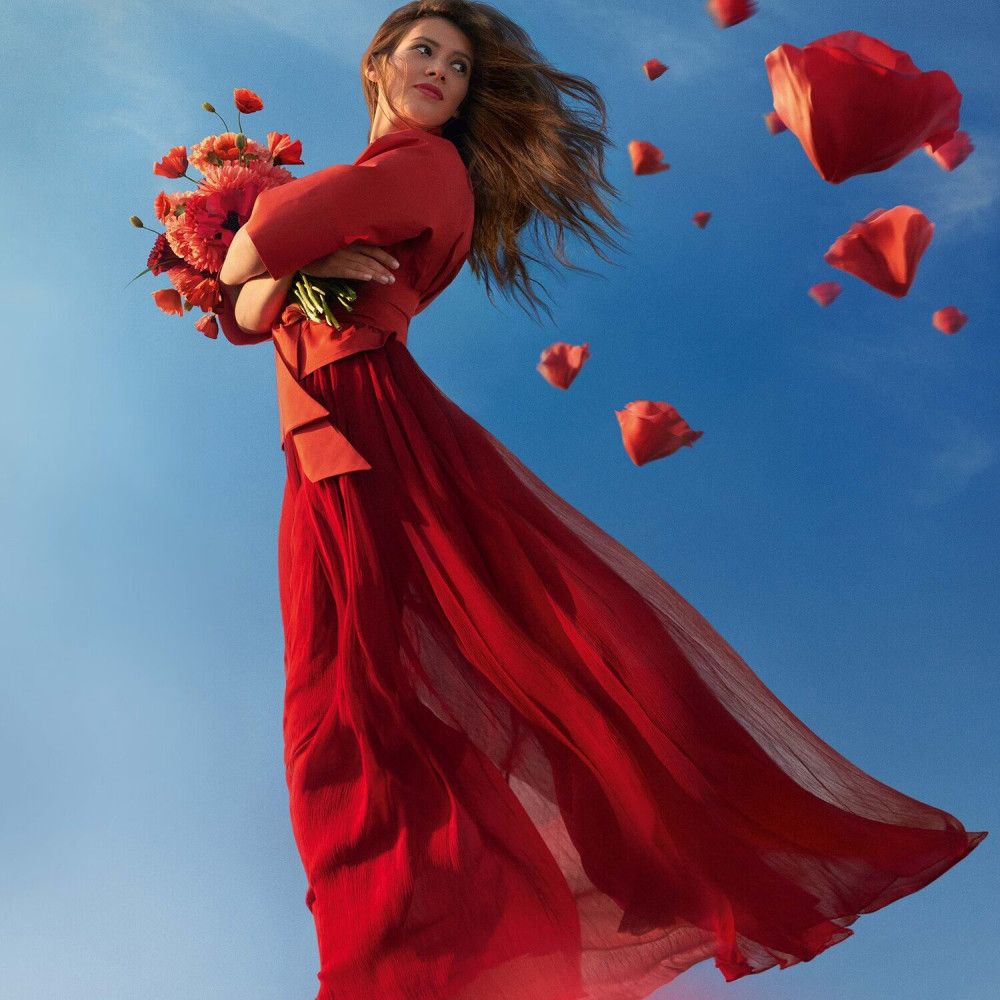Femme en robe rouge volant avec un bouquet, entourée de pétales rouges.