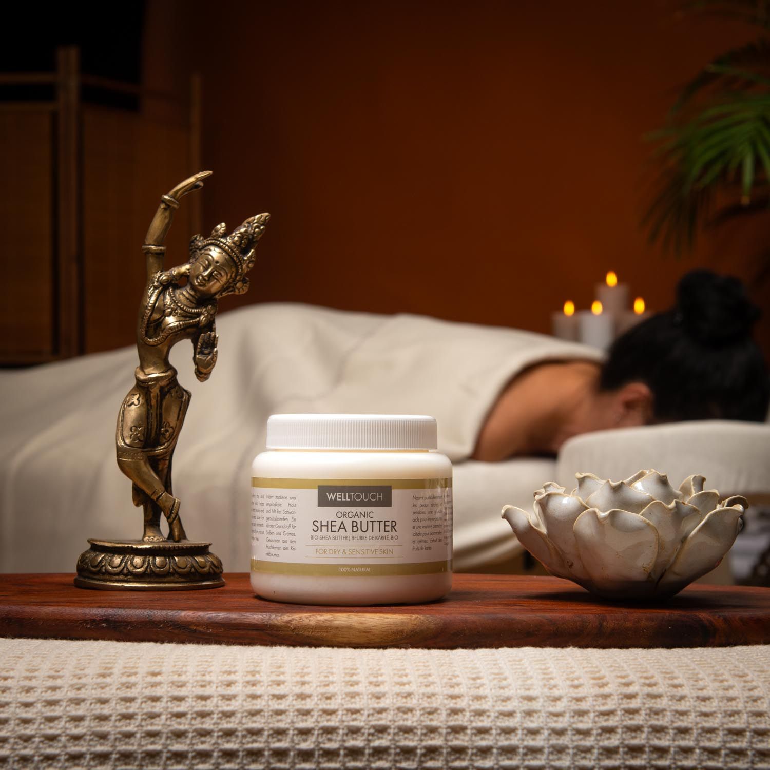 Witte pot Welltouch sheaboter. Op houten dienblad, naast gouden beeldje en lotusbloem. Persoon op massagetafel op de achtergrond.