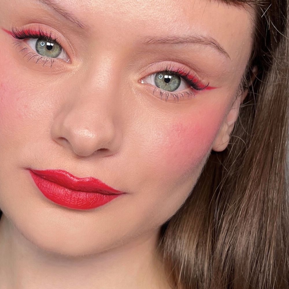 Close-up van een gezicht met rode eyeliner, blush en lippenstift. Groene ogen. Product: ALL TIGERS.