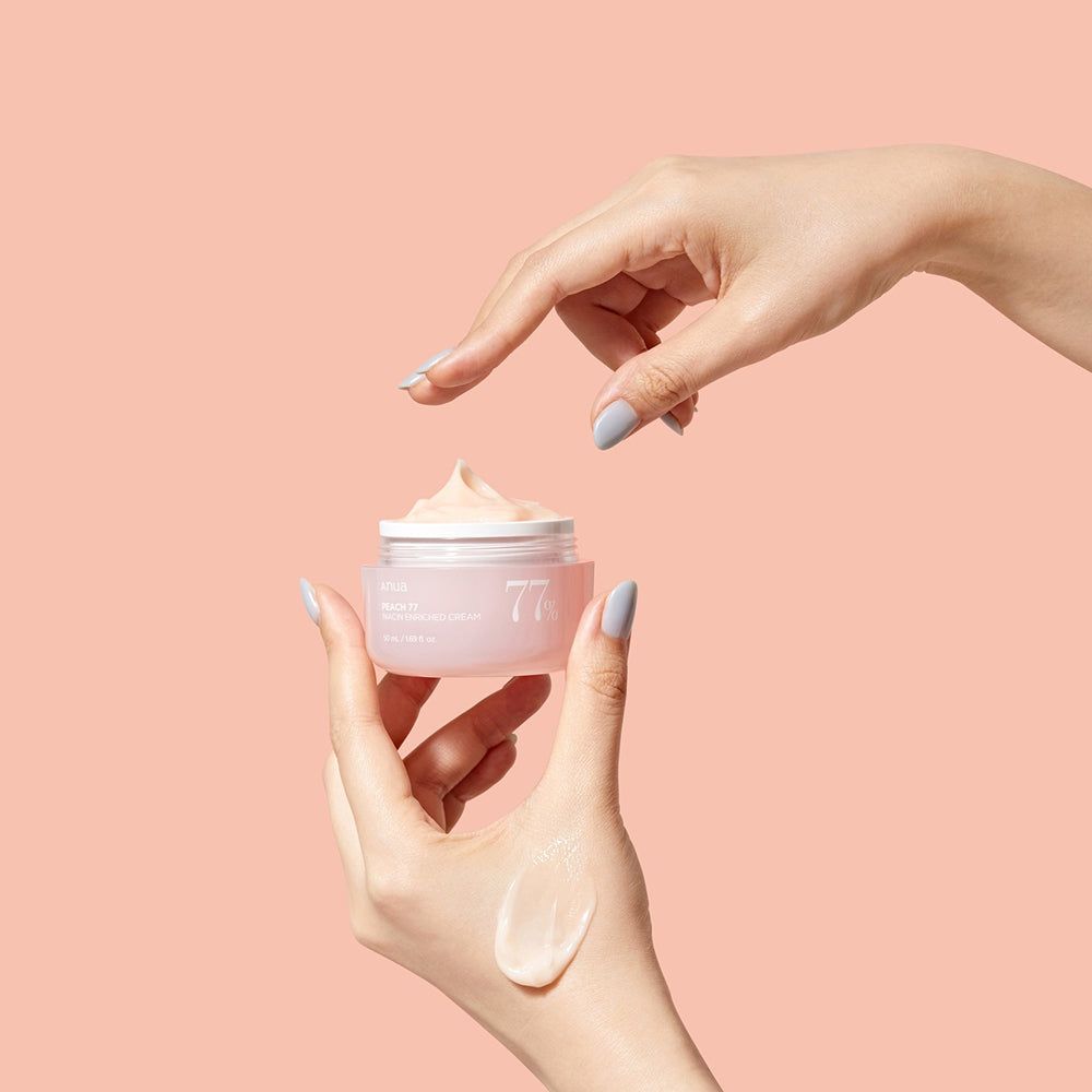 Handen die een roze crème pot vasthouden en aanraken. Opschrift: Anua, PEACH 77, Niacin Enriched Cream, 77%.