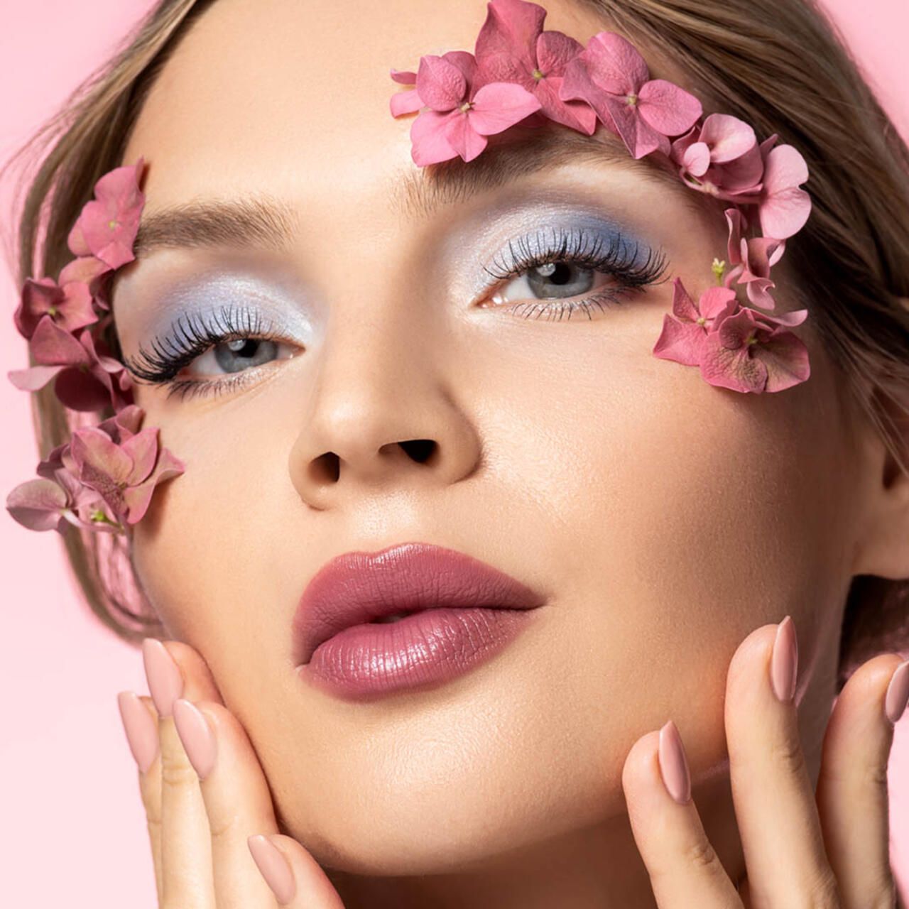 Portret van een vrouw met blauwe oogschaduw en roze lippenstift. Roze bloemen omlijsten de ogen. Lichte huid en nagels.