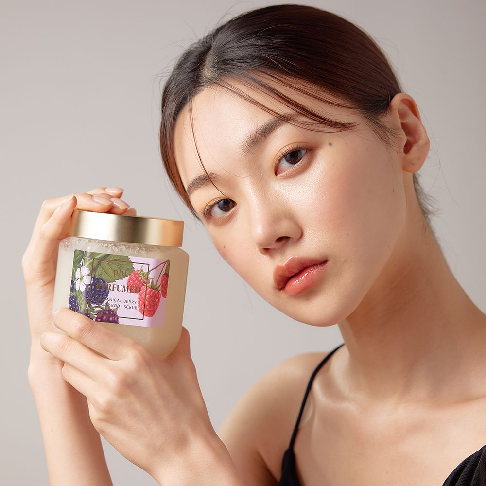 Vrouw houdt een glazen pot vast. Etiket met bessen- en bloemenillustraties. Opschrift: PERFUMED BOTANICAL BERRY SUGAR BODY SCRUB.