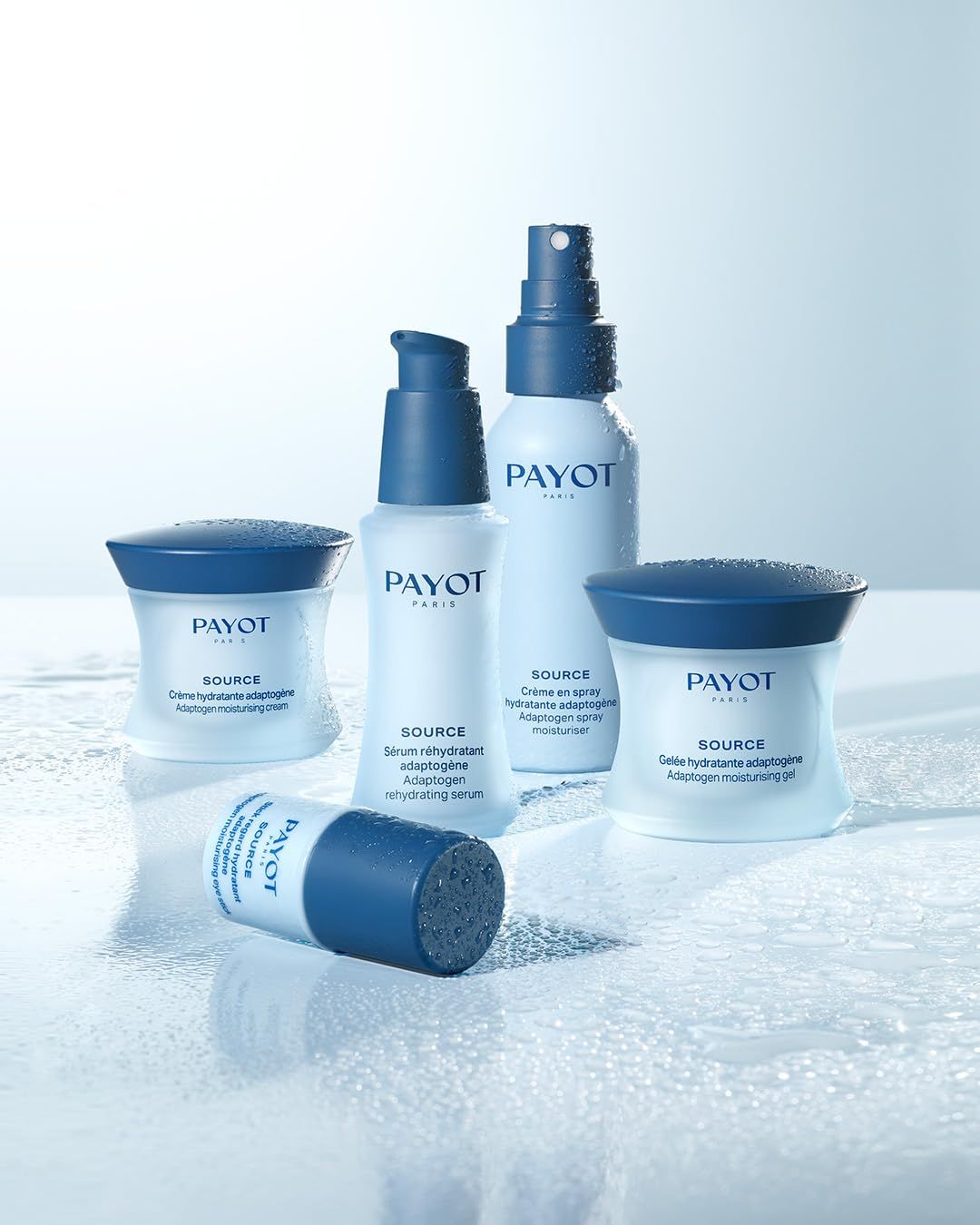 Verschillende blauwe en witte cosmetische producten, waaronder een sprayfles, op een nat oppervlak. Opschrift: PAYOT, SOURCE, Serum réhydratant, Adaptogen, moisturising serum.