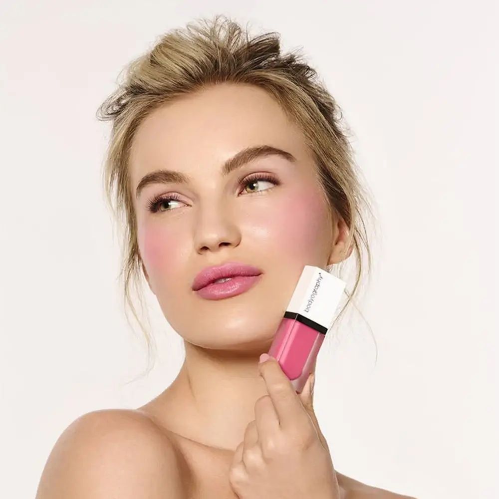 Vrouw houdt BODYOGRAPHY vloeibare blush vast. Roze blush op wangen en lippen. Witte dop, transparante houder.